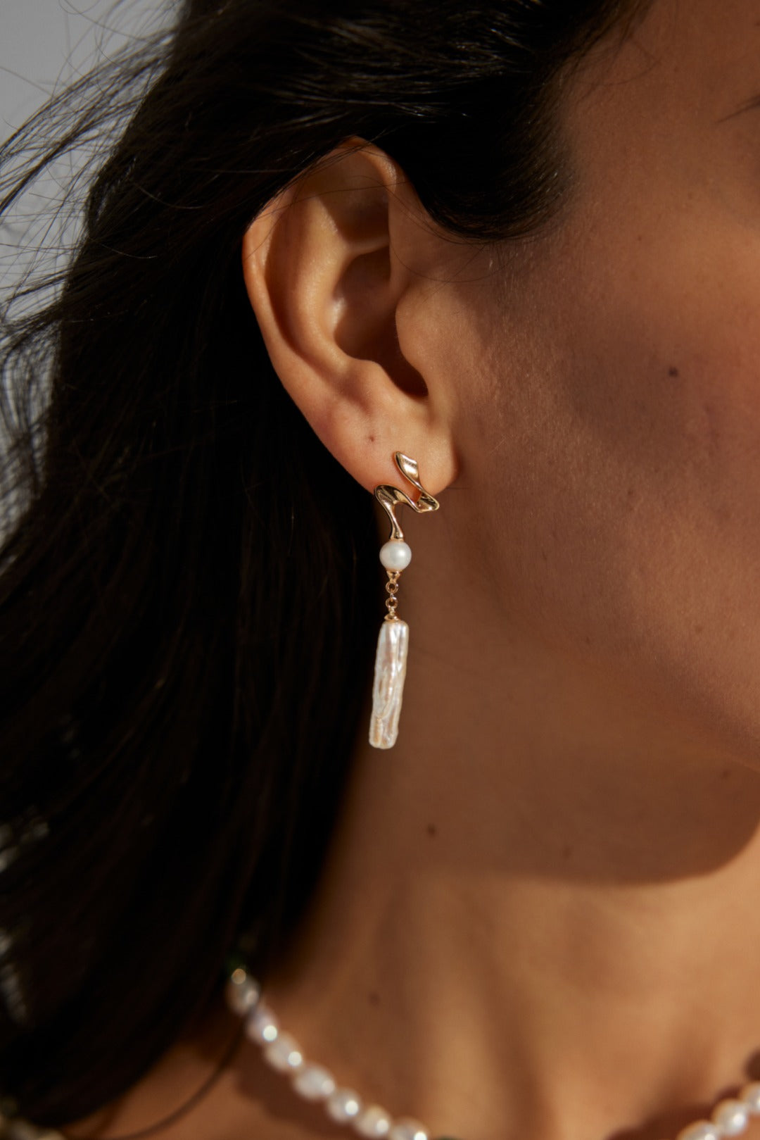 Close-up view of abstract wave baroque pearl earrings on a model's ear, featuring a gold swirl design with a dangling pearl pendant