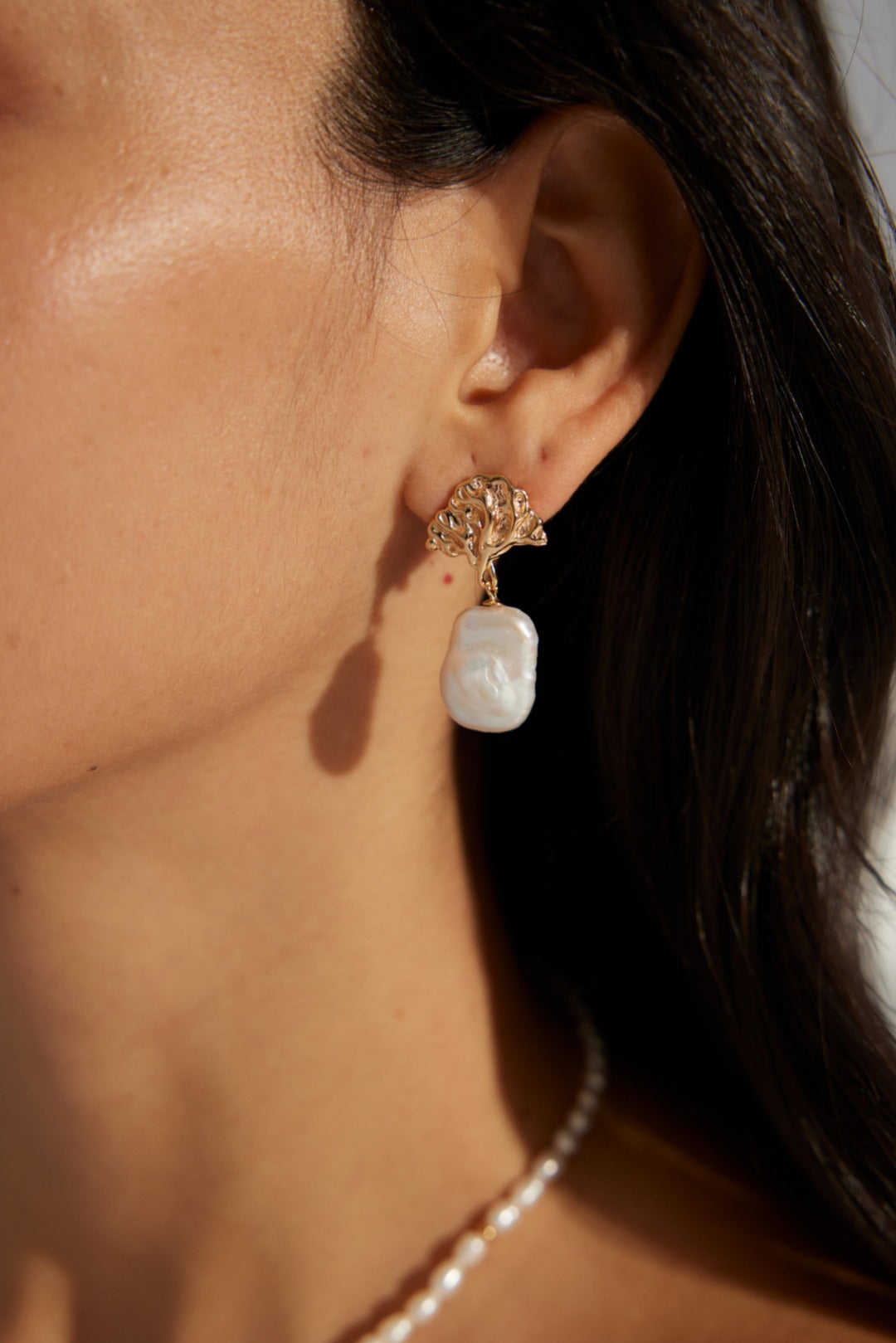 Close-up of a model wearing gold leaf-inspired baroque pearl earrings, paired with flowing dark hair and a subtle pearl necklace